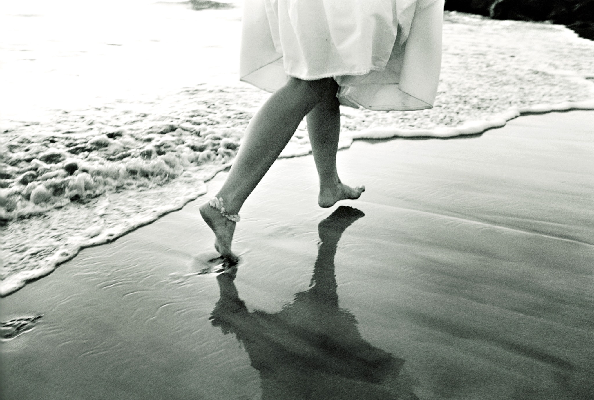 Black-and-white photograph of a barefoot bride stepping along the shoreline, her reflection falling across the wet sand Black-and-white photograph of a barefoot bride stepping along the shoreline, her reflection falling across the wet sand