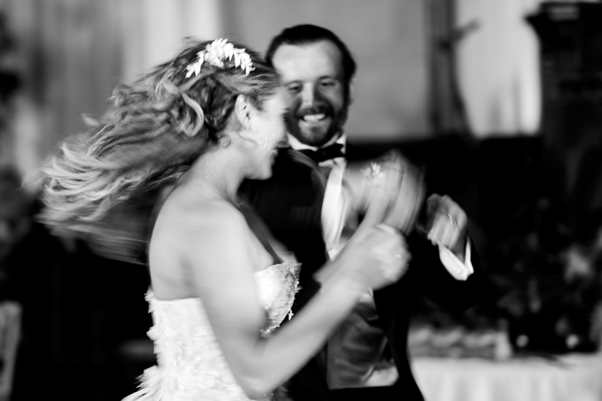A black-and-white photograph of a smiling couple dancing, with movement in the bride's hair