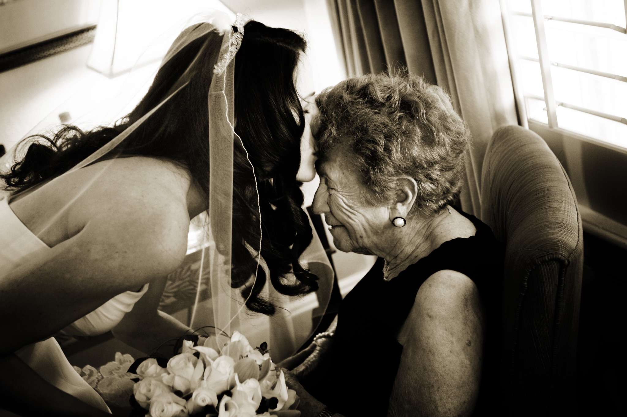A bride gently kissing an older woman's forehead beside a window